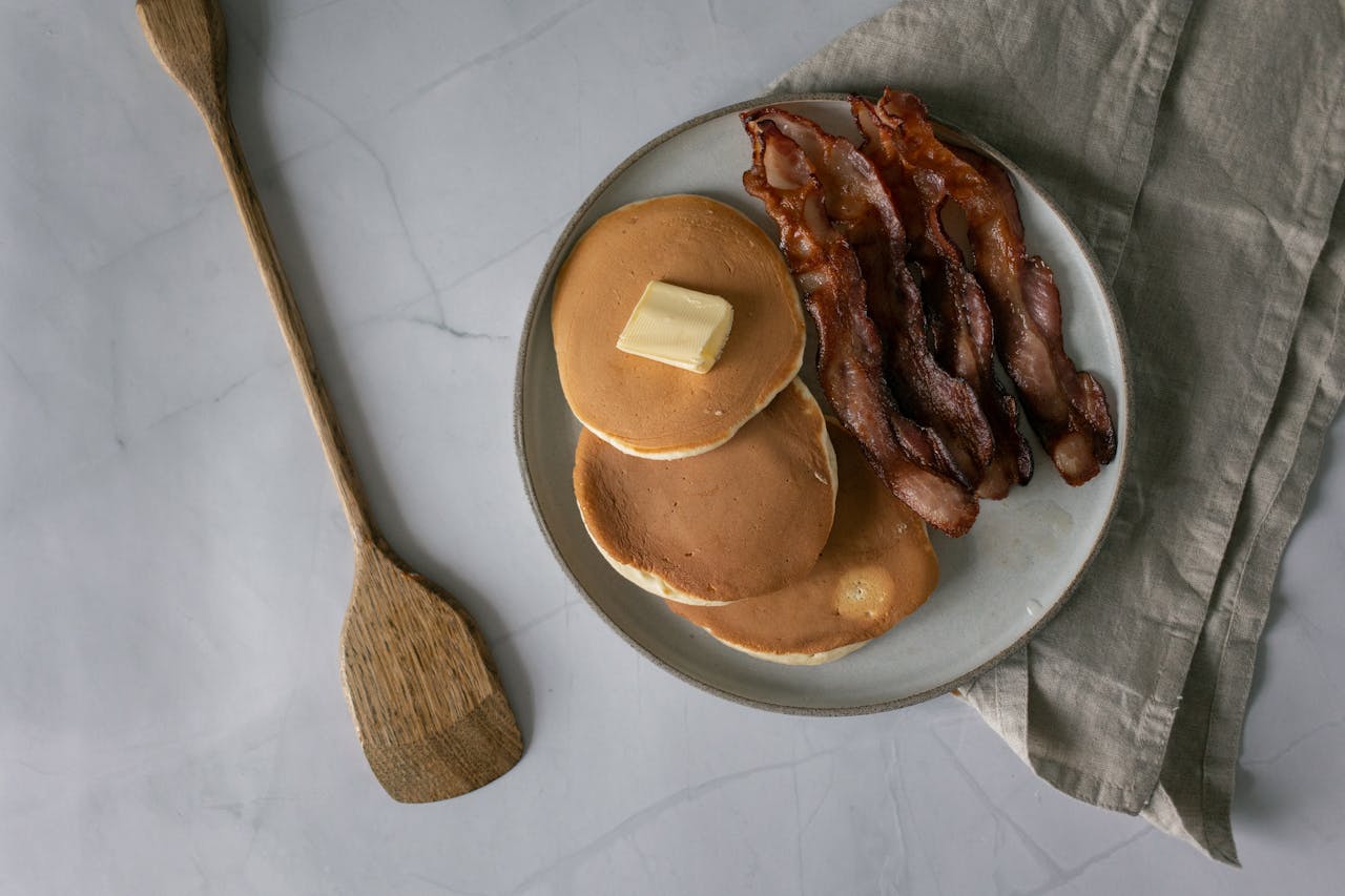 Top view of a breakfast plate with buttery pancakes and crispy bacon on a marble surface.